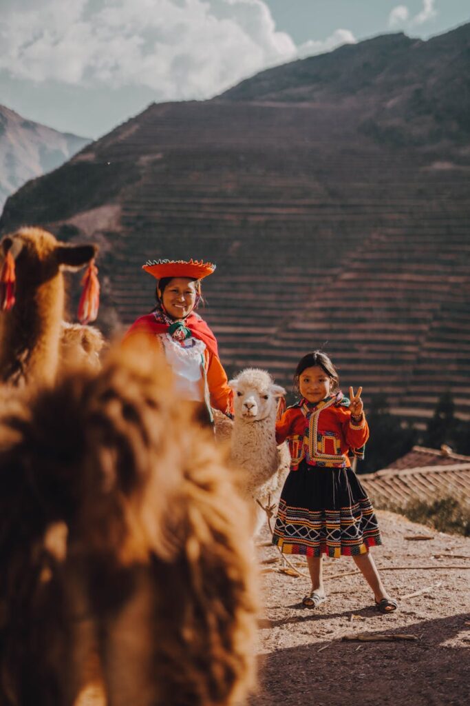 a woman and a girl in traditional clothing standing on a hill with alpacas